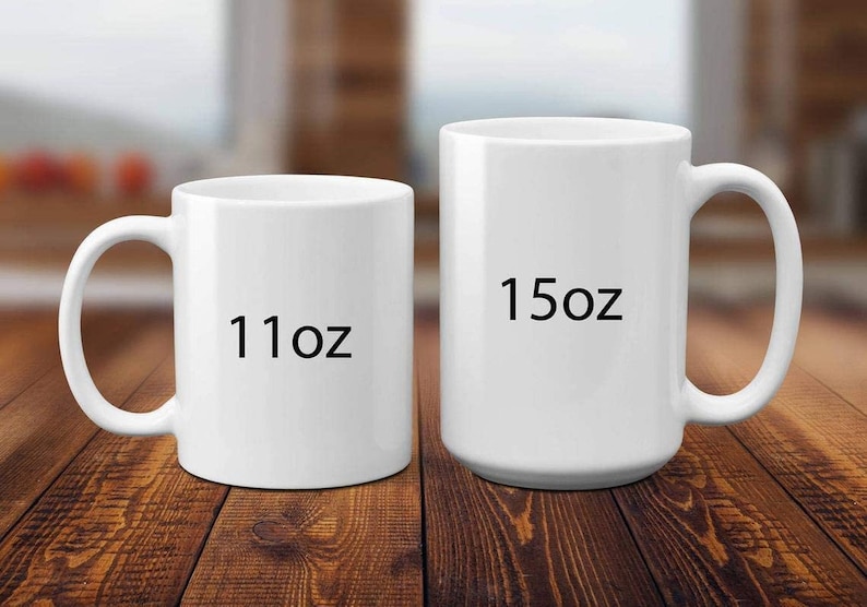 May include: Two white ceramic mugs of different sizes are displayed on a wooden surface. The smaller mug is labeled "11oz" and the larger mug is labeled "15oz". Both mugs have a curved handle.