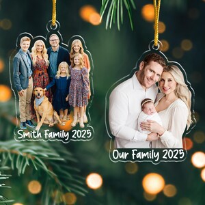 Pu&ograve; includere: Due ornamenti fotografici familiari in acrilico trasparente con cordini dorati. Uno presenta una famiglia con un cane, il testo "Smith Family 2025". L'altro mostra una famiglia con un bambino, il testo "Our Family 2025".
