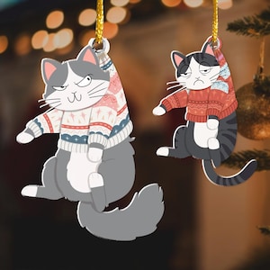 以下が含まれることがあります： 赤と白の模様のセーターを着た、グレーと白の猫のオーナメント2つ。猫は金色の紐で吊るされています。オーナメントは、クリスマスライトとクリスマスツリーのぼやけた背景に映っています。