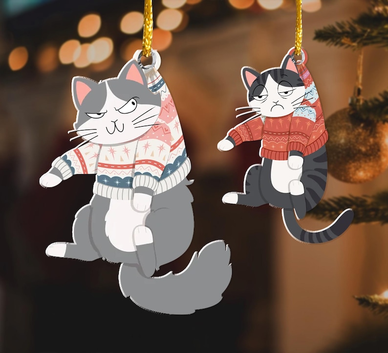 以下が含まれることがあります： 赤と白の模様のセーターを着た、グレーと白の猫のオーナメント2つ。猫は金色の紐で吊るされています。オーナメントは、クリスマスライトとクリスマスツリーのぼやけた背景に映っています。