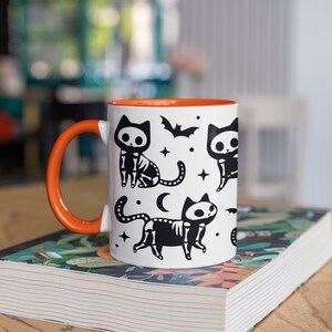 Pu&ograve; includere: Una tazza in ceramica bianca con un bordo e un manico arancioni. La tazza presenta un'illustrazione in bianco e nero di gatti scheletrici con stelle, una luna crescente e pipistrelli.