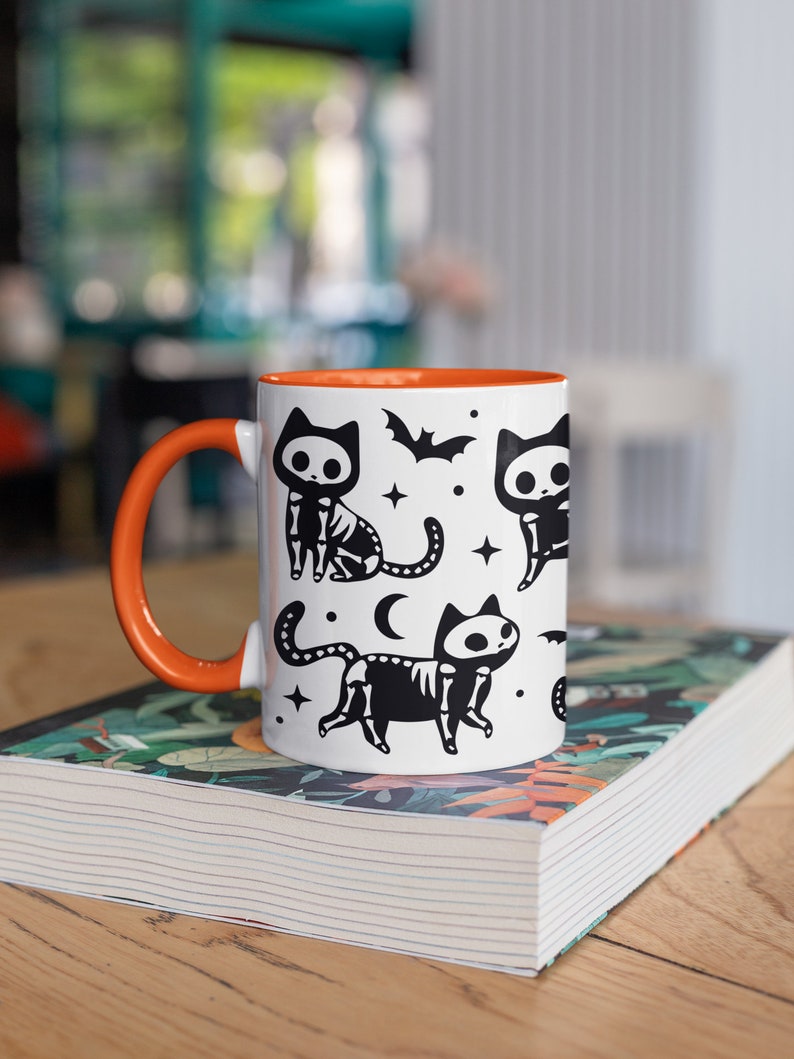 Pu&ograve; includere: Una tazza in ceramica bianca con un bordo e un manico arancioni. La tazza presenta un'illustrazione in bianco e nero di gatti scheletrici con stelle, una luna crescente e pipistrelli.