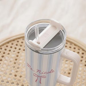 May include: A white and blue striped travel mug with a handle and a clear lid. The mug has a pink bow and the text "ms. Nicole" printed on it. A clear straw is visible.