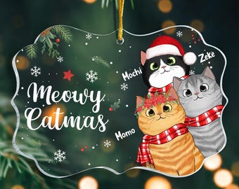 May include: A clear acrylic Christmas ornament with a white background and a festive design. It features three cats wearing Santa hats and scarves, with the text "Meowy Catmas" and the names "Momo", "Mochi", and "Zeke".