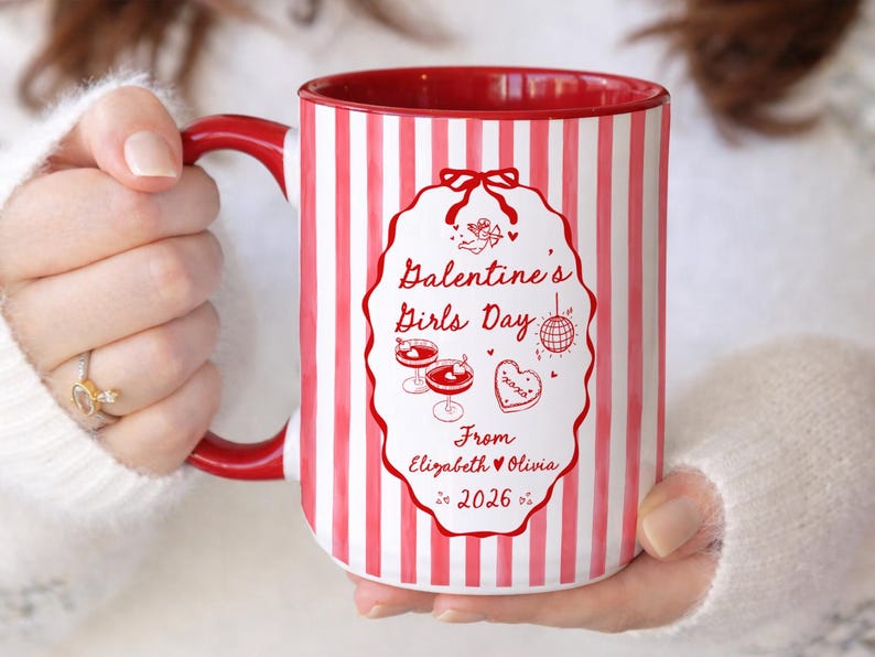 May include: A white ceramic mug with red vertical stripes and a red interior. The mug features a decorative oval with the text "Galentine's Girls Day" and illustrations of cocktails, a heart-shaped cookie, and a disco ball.