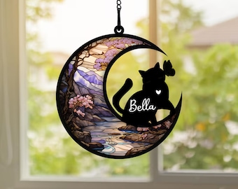 Può includere: Un acchiappasole in vetro colorato a forma di luna con una silhouette nera di un gatto seduto all'interno della luna crescente. Il gatto ha un cuore sul petto e il nome "Bella" scritto sotto. Lo sfondo dell'acchiappasole presenta un paesaggio con alberi, montagne e un fiume.