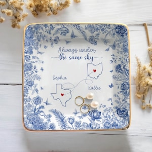May include: Square ceramic trinket dish with a blue floral design and gold trim. The dish features the text "Always under the same sky" and a map with two states and names. Two pairs of earrings are on the dish.
