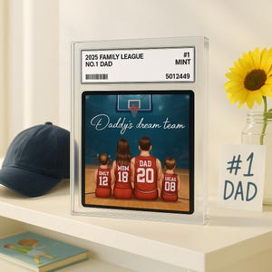 May include: A framed print featuring a family of four in basketball jerseys, with the text "Daddy's dream team." The print is encased in a clear case with the text "2025 Family League No.1 Dad." A navy blue baseball cap and a sunflower are also visible.