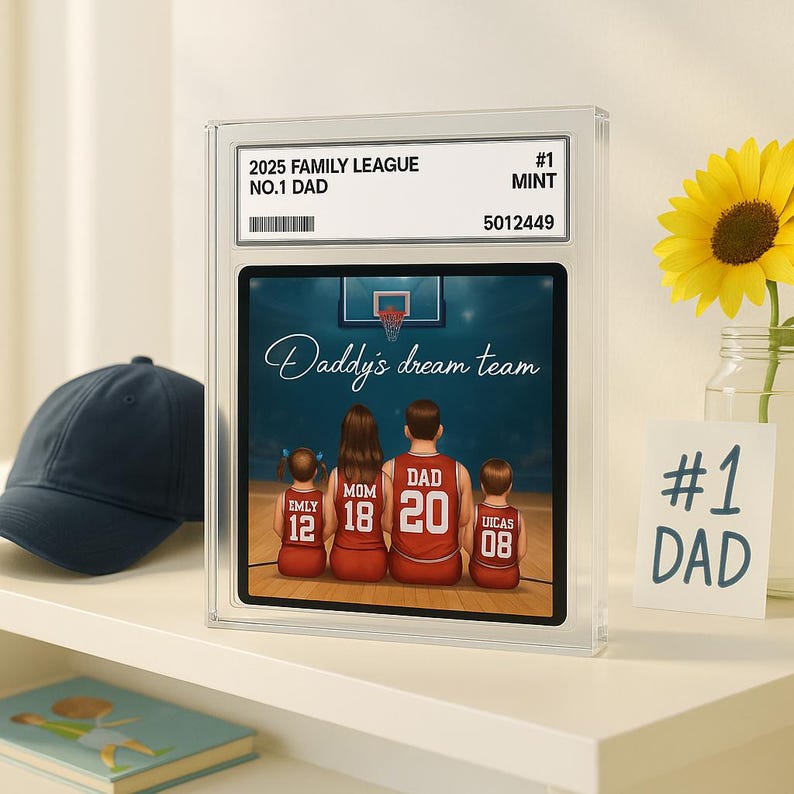 May include: A framed print featuring a family of four in basketball jerseys, with the text "Daddy's dream team." The print is encased in a clear case with the text "2025 Family League No.1 Dad." A navy blue baseball cap and a sunflower are also visible.