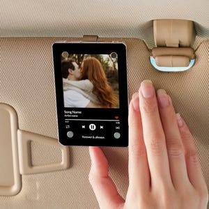 May include: A black acrylic car accessory featuring a music player interface with a photo of a couple kissing. The text "Song Name", "Artist name", and "forever & always" are visible. The accessory is clipped to a car visor.
