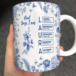 Może przedstawiać: Biały ceramiczny kubek z niebieskim kwiatowym wzorem i napisem "Bóg mówi, że jestem" oraz imieniem "LAURA". Kubek zawiera również słowa "KOCHANY", "OBFITY", "NIEZACHWIANY", "ODKUPIONY" i "OZDOBIONY".