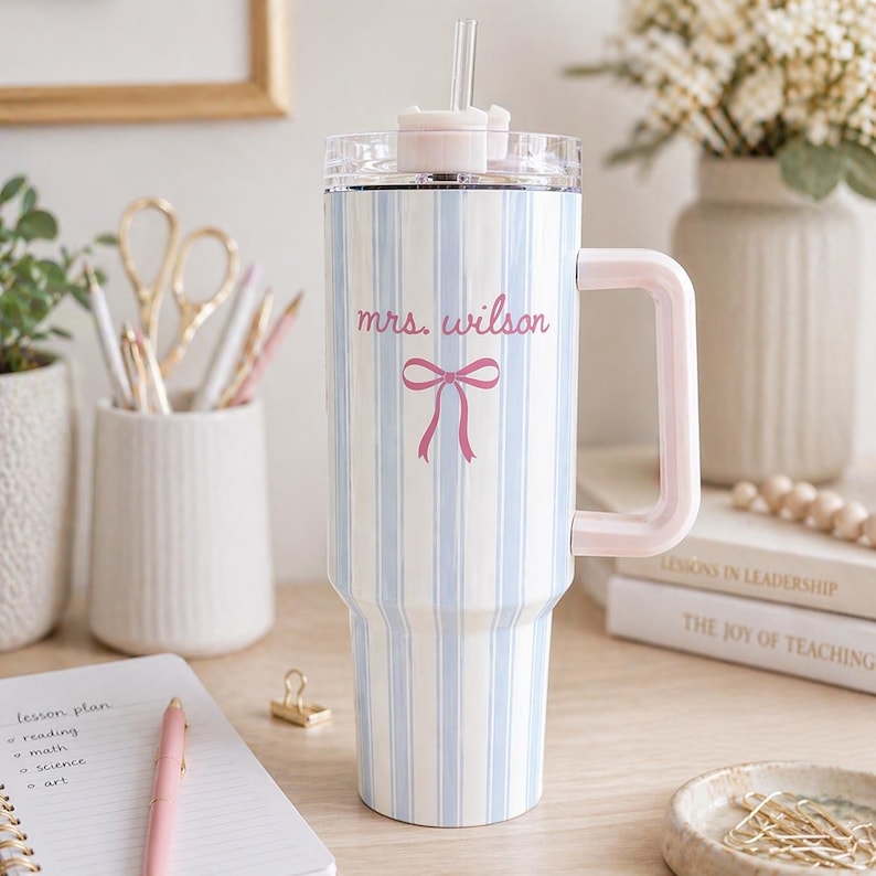 May include: A large tumbler with a light blue and white striped pattern. It has a pink handle, lid, and bow graphic with the text "mrs. wilson". Includes a clear straw. A stylish and personalized drinkware item.