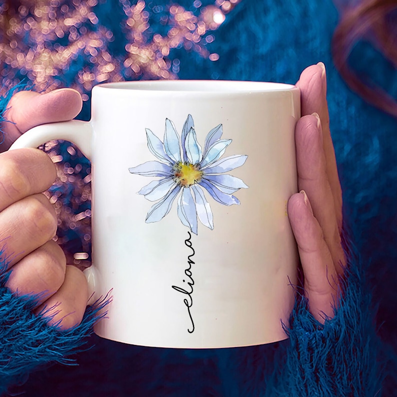 Puede incluir: Taza de cer&aacute;mica blanca con una margarita azul en acuarela y el nombre "Eliana" escrito en cursiva.