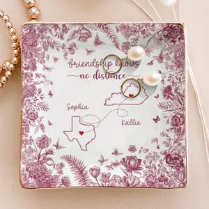 May include: Square ceramic trinket dish with a floral and butterfly design in burgundy and white. The dish features the text "friendship knows no distance" and includes a map with hearts and names. Two gold earrings with pearls rest on the dish.