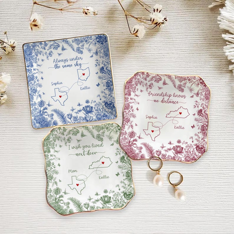 May include: Three decorative ceramic dishes with floral patterns and gold rims. Each dish features a state map with names and the phrases: "Always under the same sky", "Friendship knows no distance", and "I wish you lived next door".
