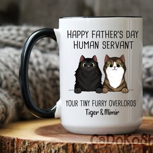 May include: White ceramic coffee mug with a black handle. The mug has a black rim and features a funny message that reads "Happy Father's Day Human Servant Your Tiny Furry Overlords Tiger & Mimir". The mug has a cartoon image of two cats.