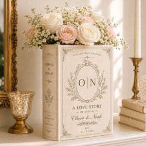Puede incluir: Una caja rectangular en forma de libro de color crema con el texto "OUR GREATEST CHAPTER", "O|N", "A LOVE STORY", "WRITTEN BY OLIVIA & NOAH", "EST. 10.12.2026", "OLIVIA & NOAH" y "THE BEGINNING OF FOREVER" con detalles florales.