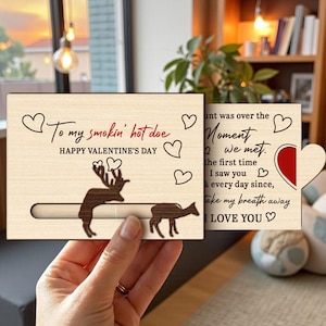 Könnte beinhalten: Eine hölzerne Valentinstagskarte mit einer Hirschsilhouette und dem Text "To my smokin' hot doe" und "Happy Valentine's Day." Die Karte lässt sich öffnen, um eine Liebesbotschaft zu enthüllen. Die Karte wird von einer Person gehalten.