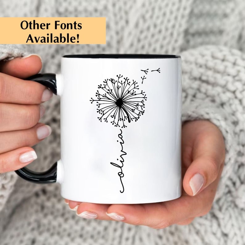 May include: White ceramic mug with a black handle. The mug has a black rim and features a black and white illustration of a dandelion with the word "olivia" written vertically below it.