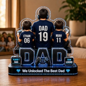 Può includere: Una luce notturna a LED personalizzata con un padre e due figli con cuffie e controller di gioco. Le magliette riportano "DAD 19", "ETHAN 06" e "NOAH 11". La base recita "We Unlocked The Best Dad".