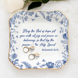 May include: A white and blue decorative ceramic tray with a gold rim, featuring a floral and butterfly design. The tray has the text "May the God of hope fill you with all joy and peace in believing, so that by the power of the Holy Spirit ROMANS 15:13". Two gold earrings with pearls rest on the tray.