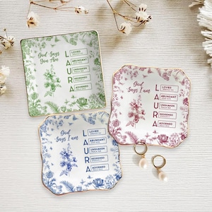 May include: Three decorative ceramic plates with floral designs in green, burgundy, and blue. Each plate has gold trim and the words "God Says You Are" or "God Says I Am" with the name "LAURA" and other inspirational words.