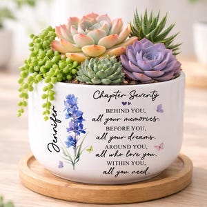 May include: A white ceramic planter with a variety of succulents, including a pink and green one. The planter has the name "Jennifer" and the words "Chapter Seventy" with a poem. It sits on a wooden tray.