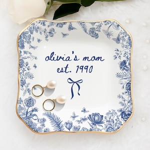 May include: A square, white ceramic trinket dish with a blue floral design and gold trim. The dish is inscribed with "olivia's mom est. 1990" and features a bow illustration. Two gold earrings with pearl drops rest on the dish.