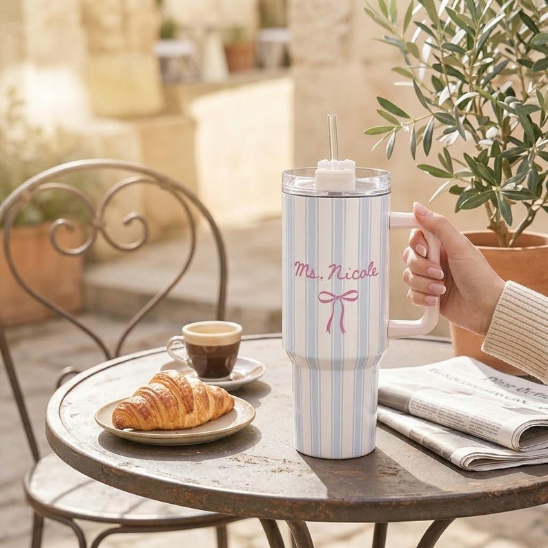 May include: A white tumbler with blue vertical stripes and a pink handle. The tumbler has the name "Ms. Nicole" in pink script with a pink bow. A croissant and coffee cup sit on a table.