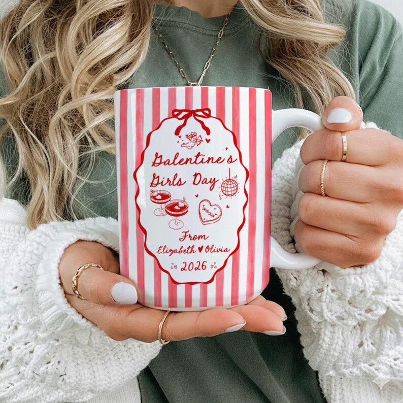 May include: A white ceramic mug with vertical red stripes. The mug features a red decorative frame with the text "Galentine's Girls Day" and other text. The mug has a white handle and is held by a person.