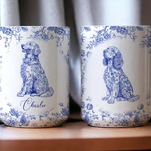 Op de afbeelding: Twee witte keramische mokken met blauwe bloemen- en hondenillustraties. Eén mok toont een hond genaamd "Charlie". De mokken hebben een klassiek ontwerp met een blauw-witte kleurstelling.