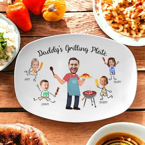 Op de afbeelding: Witte ovale serveerschaal met de tekst "Daddy's Grilling Plate" in zwart. De schaal heeft een cartoonillustratie van een familie die barbecuet, waarbij het gezicht van elk gezinslid is vervangen door een foto. De namen "Amy", "Jimmy", "Bobby" en "Mary" staan onder de figuren.