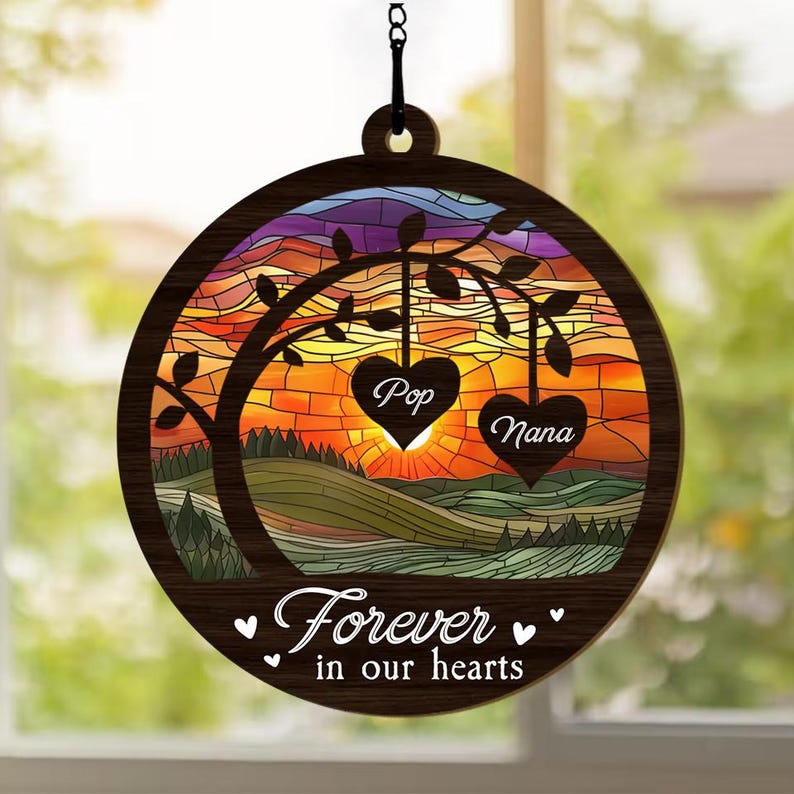 May include: A wooden suncatcher with a stained glass design of a tree with two hearts hanging from the branches. The hearts are labeled "Pop" and "Nana". The suncatcher has the text "Forever in our hearts" at the bottom.