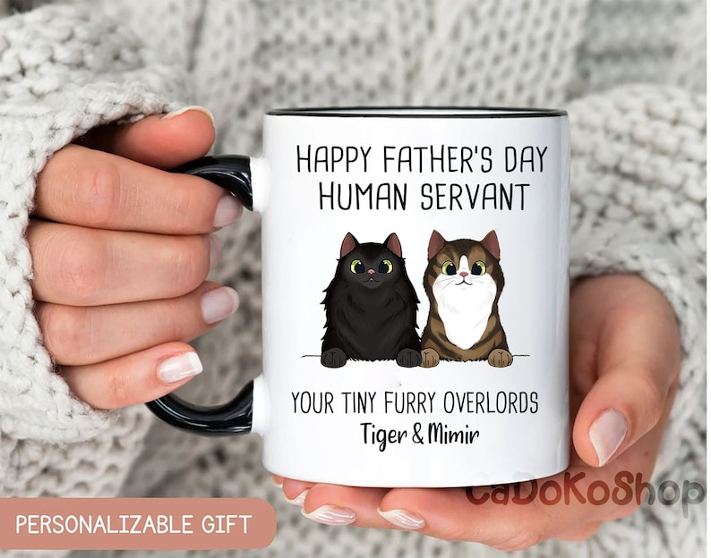 May include: White ceramic mug with a black rim and handle. The mug has a black and white illustration of two cats and the text "Happy Father's Day Human Servant Your Tiny Furry Overlords Tiger & Mimir".