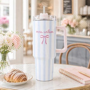 May include: A large, insulated tumbler with a light blue and white striped pattern. It has a pink handle, lid, and straw. The text "mrs. wilson" and a pink bow are printed on the front. The tumbler is on a table with a croissant and flowers.