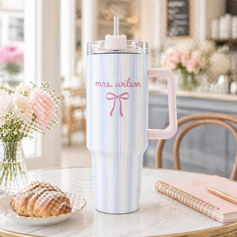 May include: A large, insulated tumbler with a light blue and white striped pattern. It has a pink handle, lid, and straw. The text "mrs. wilson" and a pink bow are printed on the front. The tumbler is on a table with a croissant and flowers.