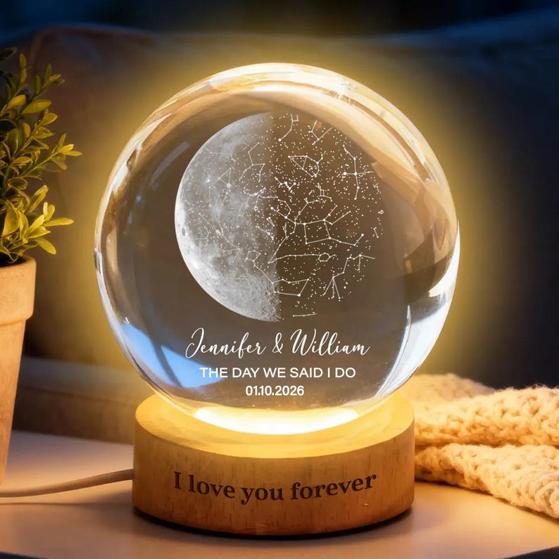 Puede incluir: Una esfera de cristal transparente con un dise&ntilde;o de luna y constelaciones, iluminada por una luz c&aacute;lida. La esfera se encuentra sobre una base de madera con el texto "I love you forever". La esfera tambi&eacute;n tiene los nombres "Jennifer & William" y la fecha "01.10.2026".