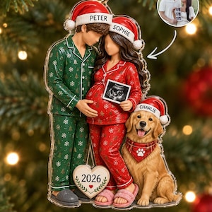 Op de afbeelding: Een doorzichtige acryl kerstornament met een koppel in groene en rode pyjama's met sneeuwvlokken en kerstmutsen met de namen Peter en Sophie. De zwangere persoon houdt een echo vast. Een golden retriever genaamd Charlie zit ernaast.