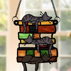 May include: A stained glass window hanging features three gray and black cats sleeping on a stack of colorful books. The cats are in various positions, with one cat on top of the stack and two cats on the lower levels. The books are stacked in a staggered pattern, with the top book being the largest and the bottom book being the smallest.