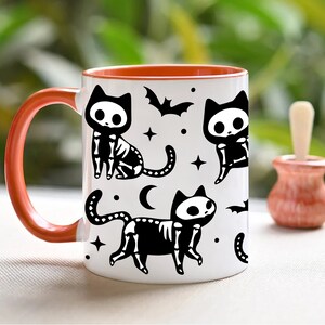 Pu&ograve; includere: Una tazza da caff&egrave; in ceramica bianca con un bordo arancione presenta un design in bianco e nero di gatti scheletrici con pipistrelli e stelle. La tazza ha un grande manico.