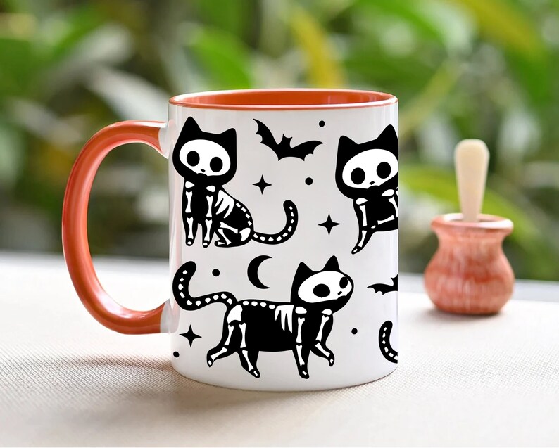 Pu&ograve; includere: Una tazza da caff&egrave; in ceramica bianca con un bordo arancione presenta un design in bianco e nero di gatti scheletrici con pipistrelli e stelle. La tazza ha un grande manico.