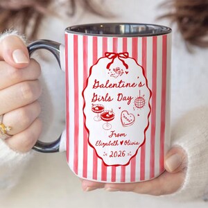 May include: A white ceramic mug with red vertical stripes and a black handle. The mug features a red oval design with the text "Galentine's Girls Day" and illustrations of cocktails, a disco ball, and a heart. The text "From Elizabeth & Olivia 2026" is also included.