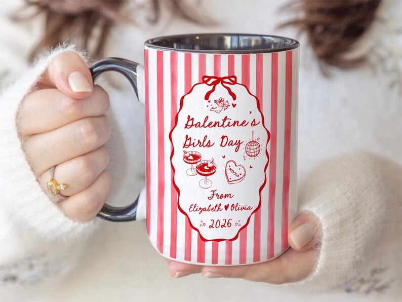 May include: A white ceramic mug with red vertical stripes and a black handle. The mug features a red oval design with the text "Galentine's Girls Day" and illustrations of cocktails, a disco ball, and a heart. The text "From Elizabeth & Olivia 2026" is also included.