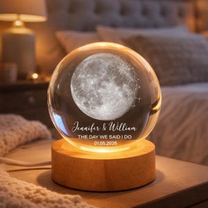 Puede incluir: Una esfera de vidrio transparente con una imagen detallada de la luna, sobre una base de madera. La esfera est&aacute; inscrita con "Jennifer & William" y "THE DAY WE SAID I DO 01.05.2026". La base emite una luz ambiental c&aacute;lida.