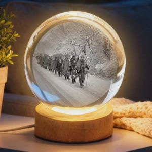 May include: A clear glass snow globe with a black and white image of people walking on a snowy road. The globe sits on a round wooden base that emits a warm, soft glow. A decorative item.