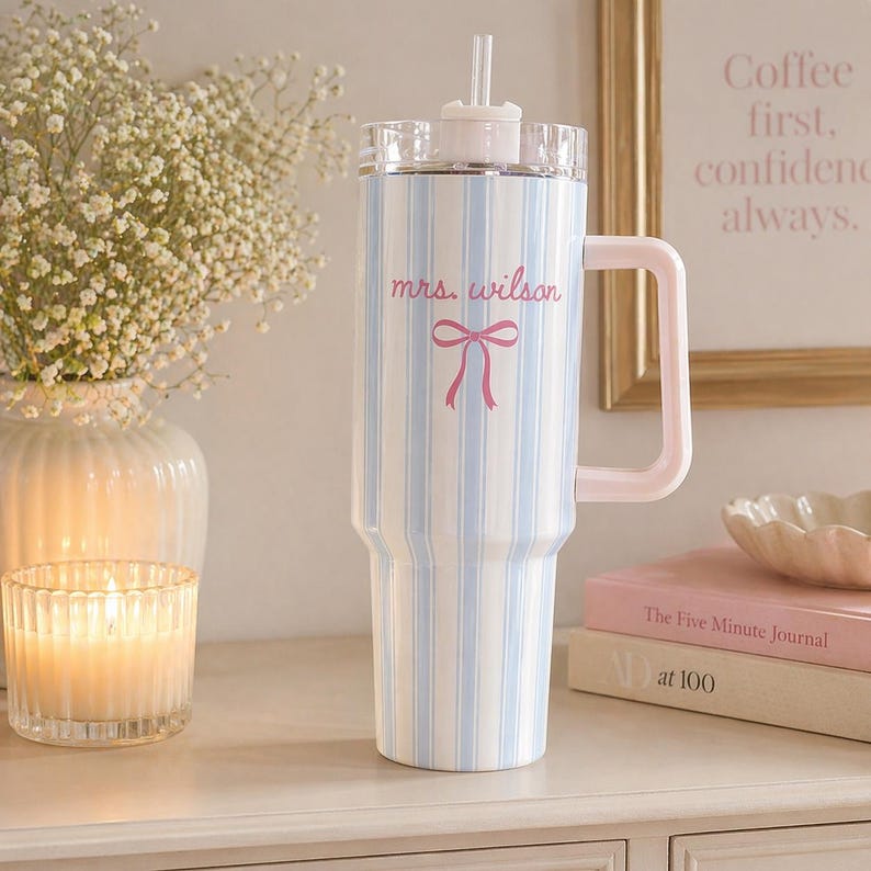 May include: A large tumbler with a light blue and white striped pattern, a pink handle, and a clear lid. The tumbler has the text "mrs. wilson" and a pink bow. A straw extends from the lid. The tumbler is next to a candle and a vase of flowers.