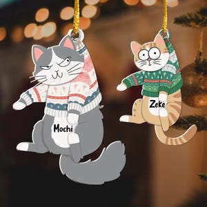 以下が含まれることがあります： セーターを着た猫の形をしたクリスマスオーナメントが2つ。灰色の猫はピンクと白のセーターを着ており、「モチ」という名前が書かれています。オレンジ色の猫は緑と白のセーターを着ており、「ゼイク」という名前が書かれています。