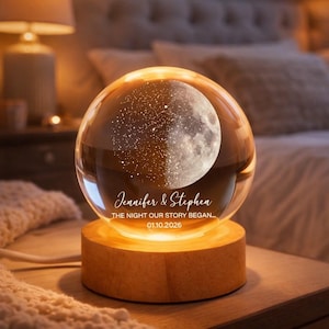 Puede incluir: Una l&aacute;mpara de noche personalizada con forma de bola de cristal y dise&ntilde;o de luna. El texto "Jennifer & Stephen" y "THE NIGHT OUR STORY BEGAN... 01.10.2026" est&aacute;n en la parte delantera. Se apoya en una base de madera.