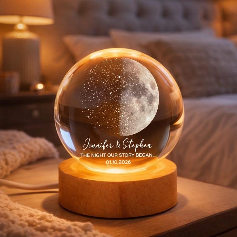 Puede incluir: Una l&aacute;mpara de noche personalizada con forma de bola de cristal y dise&ntilde;o de luna. El texto "Jennifer & Stephen" y "THE NIGHT OUR STORY BEGAN... 01.10.2026" est&aacute;n en la parte delantera. Se apoya en una base de madera.