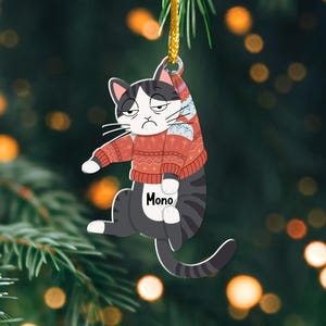 以下が含まれることがあります： 「Mono」と印刷された赤と白のセーターを着た白黒の猫。猫は金色の紐にぶら下がっていて、クリスマスのオーナメントです。